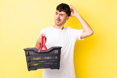 Genç İspanyol adam şaşkın ve kafası karışık, kaşınan bir kafa hissediyor. boş alışveriş sepeti kavramı