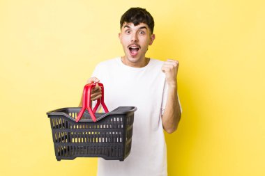 Genç İspanyol adam şok olmuş hissediyor, gülüyor ve başarıyı kutluyor. boş alışveriş sepeti kavramı