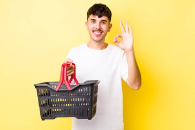 Genç İspanyol adam kendini mutlu hissediyor, iyi bir jestle onaylıyor. boş alışveriş sepeti kavramı