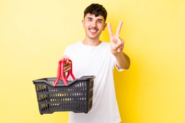 Genç İspanyol adam gülümsüyor ve mutlu görünüyor, zafer ya da barış işareti yapıyor. boş alışveriş sepeti kavramı