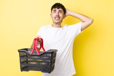 Genç İspanyol bir adam stresli, endişeli ya da korkmuş hissediyor, elleri başının üstünde. boş alışveriş sepeti kavramı