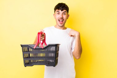 Genç İspanyol bir adam mutlu hissediyor ve heyecanla kendini işaret ediyor. boş alışveriş sepeti kavramı