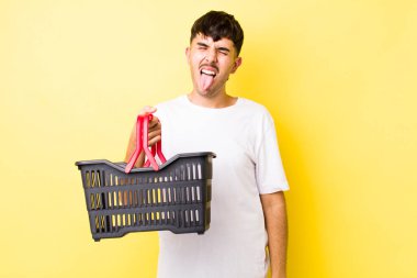 Neşeli ve asi tavırlı, şaka yapan ve dilini dışarı çıkaran genç İspanyol bir adam. boş alışveriş sepeti kavramı