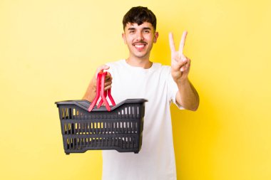 Genç İspanyol adam gülümsüyor ve arkadaş canlısı görünüyor, iki numarayı gösteriyor. boş alışveriş sepeti kavramı