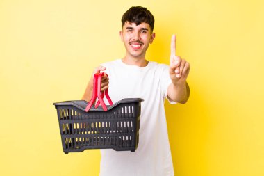 Genç İspanyol adam gülümsüyor ve arkadaş canlısı görünüyor, bir numarayı gösteriyor. boş alışveriş sepeti kavramı