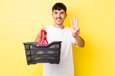 Genç İspanyol adam gülümsüyor ve arkadaş canlısı görünüyor, üç numarayı gösteriyor. boş alışveriş sepeti kavramı