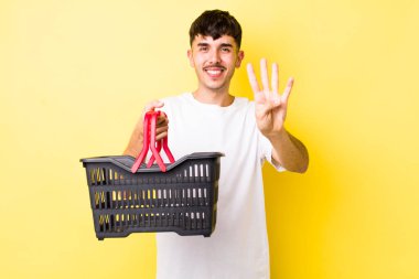 Genç İspanyol adam gülümsüyor ve arkadaş canlısı görünüyor, dört numarayı gösteriyor. boş alışveriş sepeti kavramı