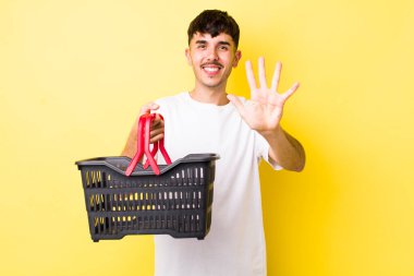 Genç İspanyol adam gülümsüyor ve arkadaş canlısı görünüyor, beş numarayı gösteriyor. boş alışveriş sepeti kavramı