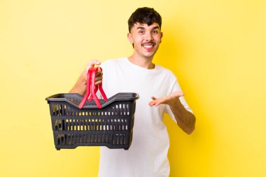 Genç İspanyol adam neşeyle gülümsüyor, mutlu hissediyor ve bir konsept gösteriyor. boş alışveriş sepeti kavramı