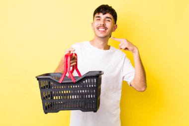 Genç İspanyol adam kendine güvenerek gülümsüyor ve kendi gülüşünü gösteriyor. boş alışveriş sepeti kavramı