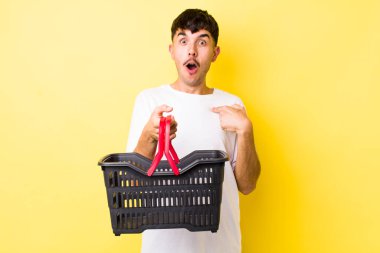 Genç İspanyol bir adam şok olmuş ve ağzı açık, kendini işaret ederek şaşırmış görünüyor. boş alışveriş sepeti kavramı