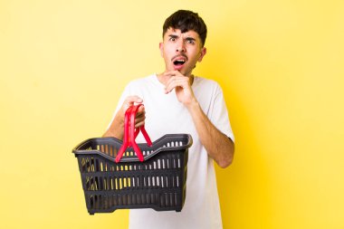 Ağzı ve gözleri sonuna kadar açık ve eli çenesinde genç İspanyol bir adam. boş alışveriş sepeti kavramı
