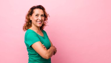 Orta yaşlı İspanyol kadın kameraya çapraz kollarıyla gülümsüyor ve mutlu, kendinden emin, tatmin olmuş bir yüz ifadesi var.