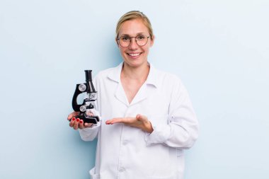 Genç sarışın kadın neşeli bir şekilde gülümsüyor, mutlu hissediyor ve bir konsept gösteriyor. Mikroskop konseptine sahip bilim adamı