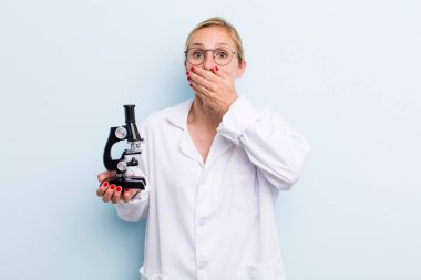 Genç, sarışın bir kadın şok olmuş bir şekilde elleriyle ağzını kapatıyor. Mikroskop konseptine sahip bilim adamı