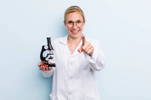 young adult blonde woman pointing at camera choosing you. scientist with a microscope concept
