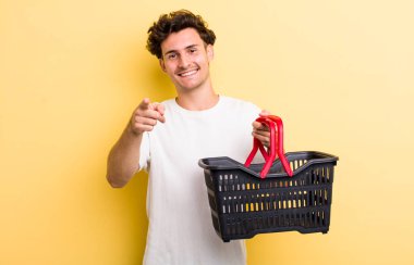 Genç ve yakışıklı bir adam kamerayı işaret ederek seni seçiyor. boş alışveriş sepeti kavramı