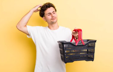 Kafası karışmış, kafası karışmış, kaşınan genç yakışıklı bir adam. boş alışveriş sepeti kavramı