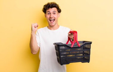 Genç yakışıklı adam şok olmuş hissediyor, gülüyor ve başarıyı kutluyor. boş alışveriş sepeti kavramı