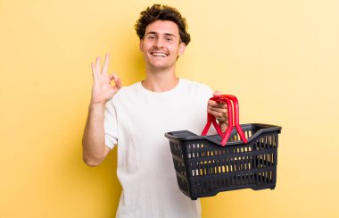 Genç yakışıklı adam kendini mutlu hissediyor, iyi bir jestle onaylıyor. boş alışveriş sepeti kavramı