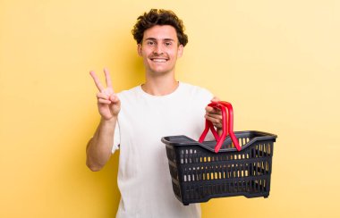 Genç yakışıklı adam gülümsüyor ve mutlu görünüyor, zafer ya da barış işareti yapıyor. boş alışveriş sepeti kavramı