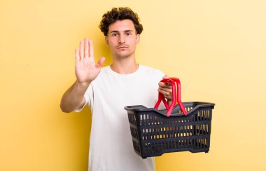 Genç ve yakışıklı adam ciddi görünüyor. Avuç içi gösteriyor. Durma hareketi yapıyor. boş alışveriş sepeti kavramı