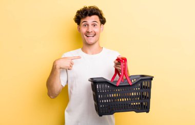 Genç yakışıklı adam kendini mutlu hissediyor ve heyecanla kendini işaret ediyor. boş alışveriş sepeti kavramı