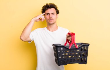 Genç yakışıklı, kafası karışık ve şaşkın hissediyor, sana deli olduğunu gösteriyor. boş alışveriş sepeti kavramı