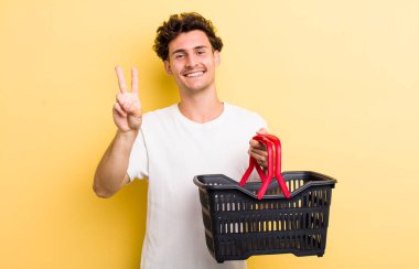 Genç yakışıklı adam gülümsüyor ve arkadaş canlısı görünüyor, iki numarayı gösteriyor. boş alışveriş sepeti kavramı