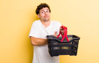 Genç yakışıklı adam omuz silkiyor, kafası karışık ve kararsız hissediyor. boş alışveriş sepeti kavramı