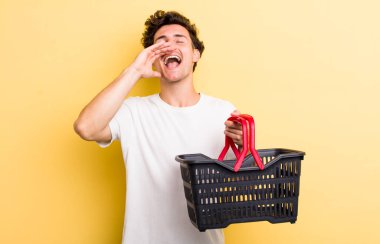 Genç yakışıklı adam kendini mutlu hissediyor, elleri ağzının kenarında bağırıyor. boş alışveriş sepeti kavramı