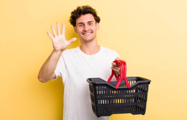 Genç yakışıklı adam gülümsüyor ve arkadaş canlısı görünüyor, beş numarayı gösteriyor. boş alışveriş sepeti kavramı