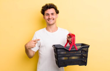 Genç yakışıklı adam neşeyle gülümsüyor, mutlu hissediyor ve bir tarafı işaret ediyor. boş alışveriş sepeti kavramı