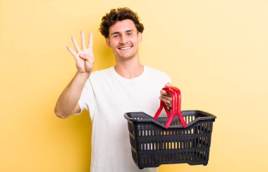 Genç yakışıklı adam gülümsüyor ve arkadaş canlısı görünüyor, dört numarayı gösteriyor. boş alışveriş sepeti kavramı