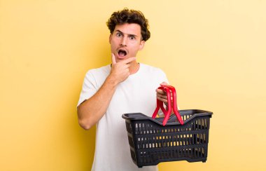 Ağzı ve gözleri sonuna kadar açık ve eli çenesinde olan yakışıklı bir genç. boş alışveriş sepeti kavramı