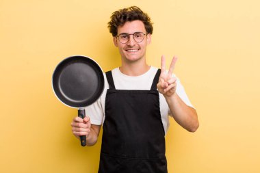 Genç yakışıklı adam gülümsüyor ve mutlu görünüyor, zafer ya da barış işareti yapıyor. Şefin tava konsepti.