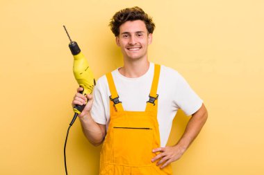 Genç yakışıklı adam mutlu bir şekilde gülümsüyor, eli kalçasında ve kendinden emin. Matkap konsepti olan bir tamirci.