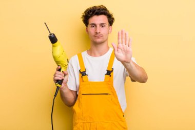 Genç ve yakışıklı adam ciddi görünüyor. Avuç içi gösteriyor. Durma hareketi yapıyor. Matkap konsepti olan bir tamirci.