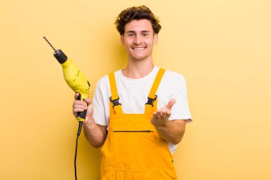 Genç yakışıklı adam mutlu bir şekilde gülümsüyor ve bir konsept sunuyor. Matkap konsepti olan bir tamirci.