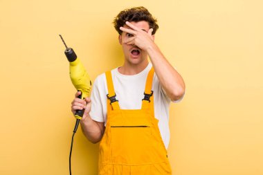 Genç, yakışıklı adam şok olmuş, korkmuş ya da korkmuş görünüyor, yüzünü eliyle kapatıyor. Matkap konsepti olan bir tamirci.