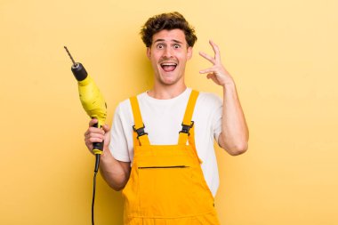 Genç yakışıklı adam ellerini havaya kaldırarak çığlık atıyor. Matkap konsepti olan bir tamirci.