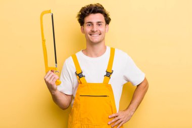 Genç yakışıklı adam mutlu bir şekilde gülümsüyor, eli kalçasında ve kendinden emin. Tamirci ve testere konsepti