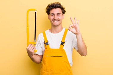 Genç yakışıklı adam kendini mutlu hissediyor, iyi bir jestle onaylıyor. Tamirci ve testere konsepti