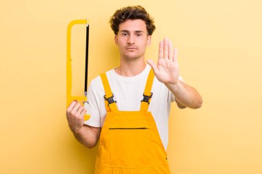 Genç ve yakışıklı adam ciddi görünüyor. Avuç içi gösteriyor. Durma hareketi yapıyor. Tamirci ve testere konsepti