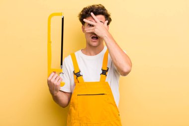 Genç, yakışıklı adam şok olmuş, korkmuş ya da korkmuş görünüyor, yüzünü eliyle kapatıyor. Tamirci ve testere konsepti