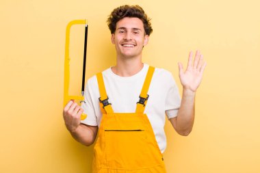 Genç yakışıklı adam mutlu bir şekilde gülümsüyor, el sallıyor, hoş geldin diyor ve seni selamlıyor. Tamirci ve testere konsepti