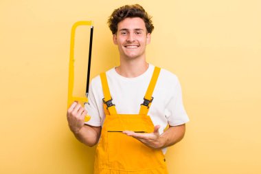 Genç yakışıklı adam neşeyle gülümsüyor, mutlu hissediyor ve bir konsept gösteriyor. Tamirci ve testere konsepti