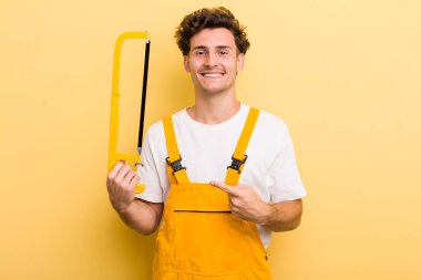 Genç yakışıklı adam neşeyle gülümsüyor, mutlu hissediyor ve bir tarafı işaret ediyor. Tamirci ve testere konsepti