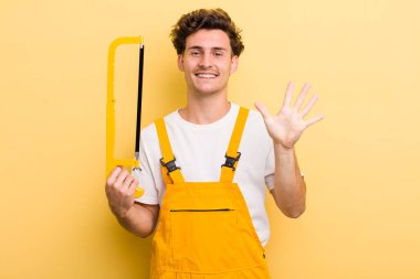 Genç yakışıklı adam gülümsüyor ve arkadaş canlısı görünüyor, beş numarayı gösteriyor. Tamirci ve testere konsepti