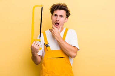 Ağzı ve gözleri sonuna kadar açık ve eli çenesinde olan yakışıklı bir genç. Tamirci ve testere konsepti